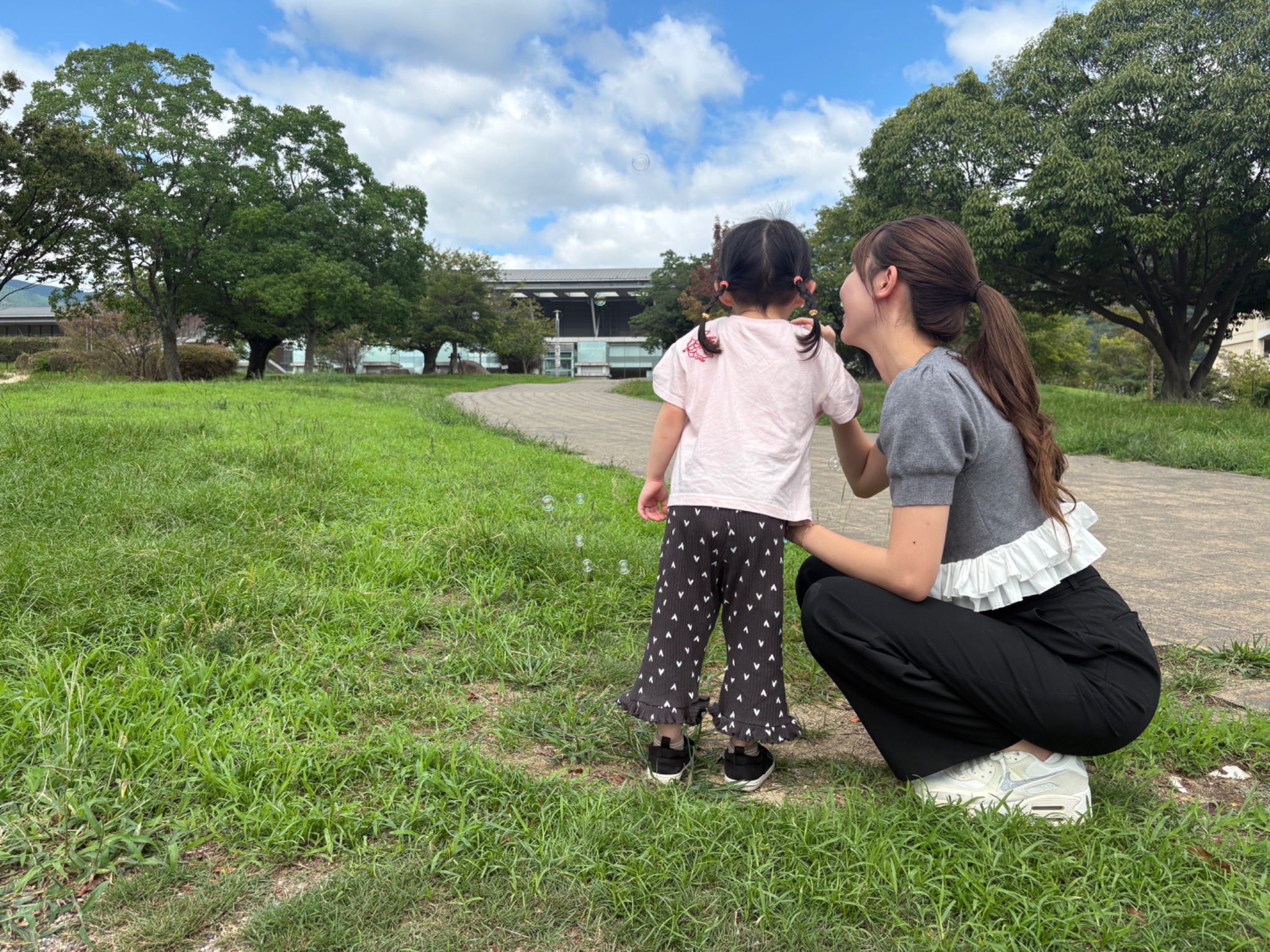 芝生の上で、シッターと子どもが一緒にシャボン玉を見ている後ろ姿